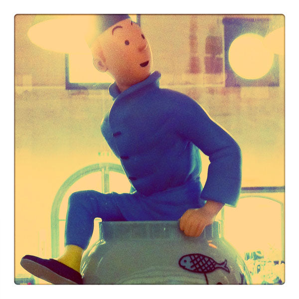 A Tintin toy in a blue outfit and hat appearing to climb out of a ceramic vase decorated with a fish design.