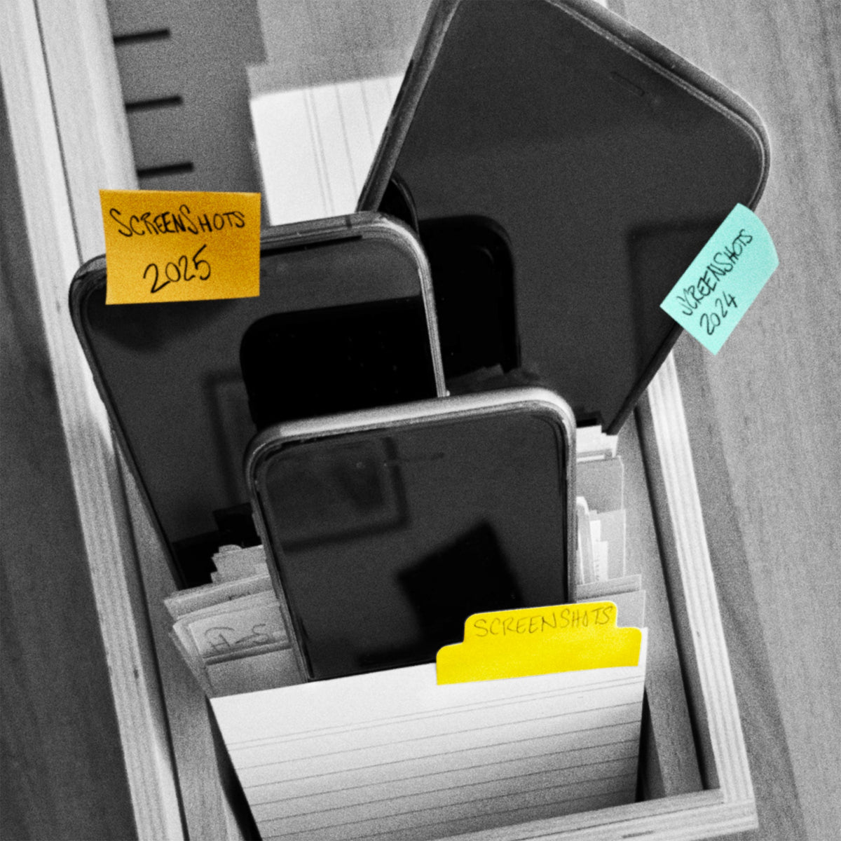 Black-and-white photo of a small wooden card catalog box with its lid open, showing a divider tab labeled SCREENSHOTS with three old iPhones filed behind it, labeled “Screenshots 2024” and “Screenshots 2025.”