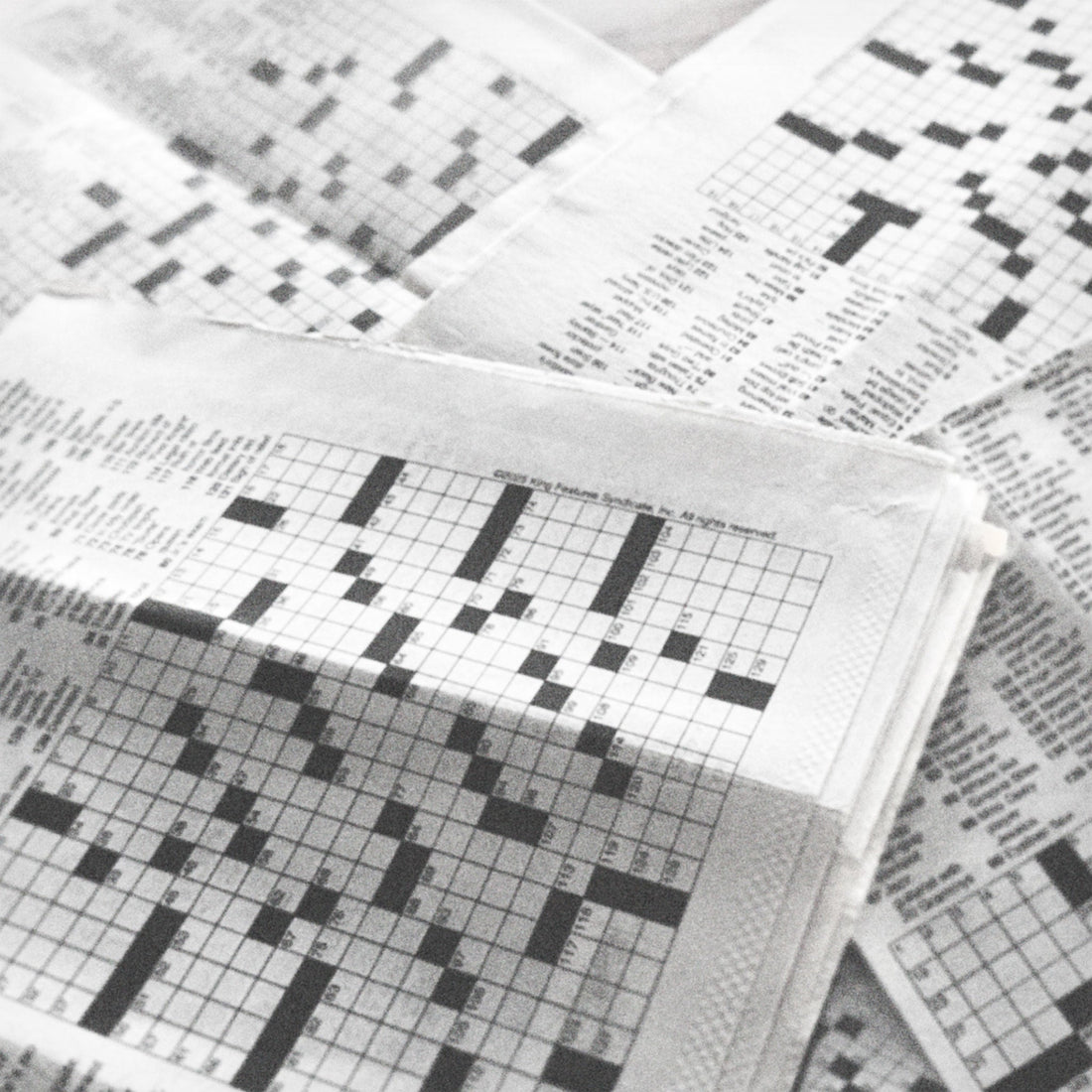 Black and white photo of multiple newspapers with crossword puzzles piled one on top of the other, with empty grids.
