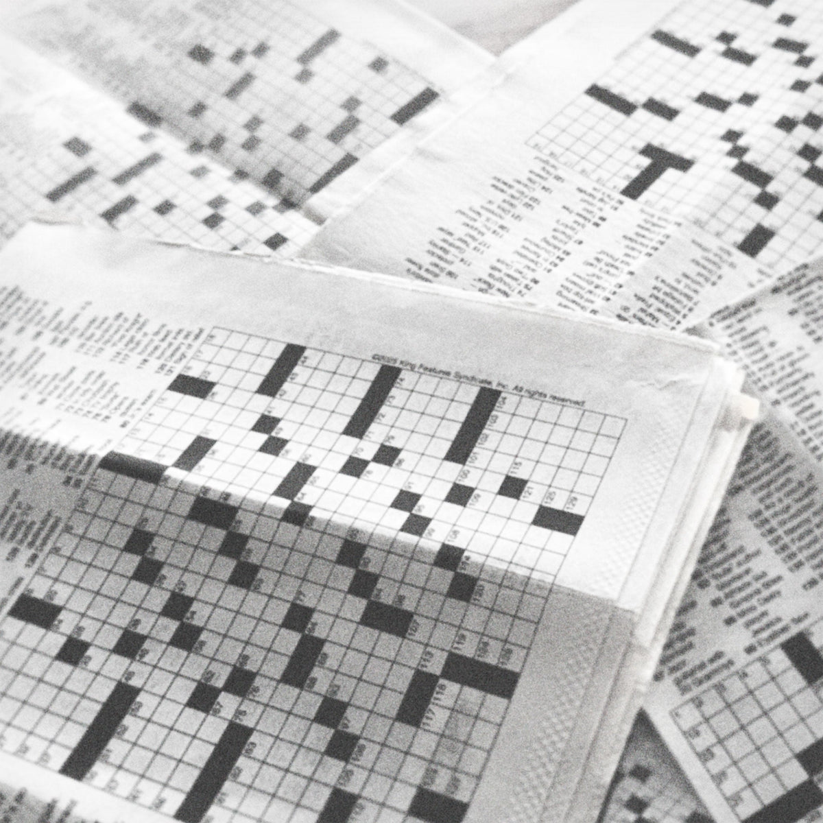 Black and white photo of multiple newspapers with crossword puzzles piled one on top of the other, with empty grids.