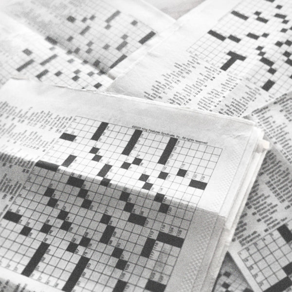 Black and white photo of multiple newspapers with crossword puzzles piled one on top of the other, with empty grids.