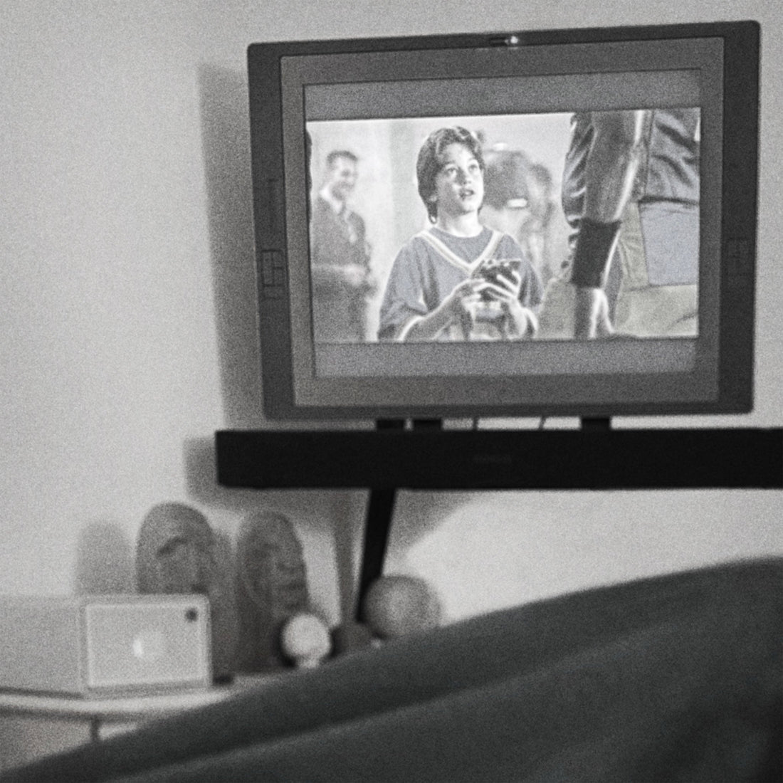 Black-and-white photo taken from under a blanket with feet up, looking at a wall-mounted monitor playing an episode of Psych above a shelf of hand-carved wooden sculptures.