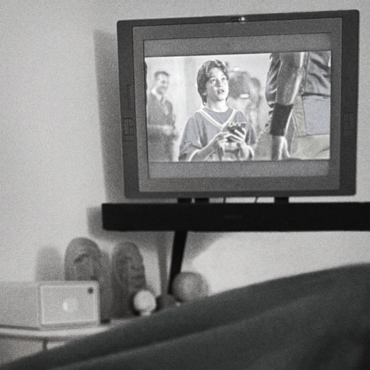 Black-and-white photo taken from under a blanket with feet up, looking at a wall-mounted monitor playing an episode of Psych above a shelf of hand-carved wooden sculptures.
