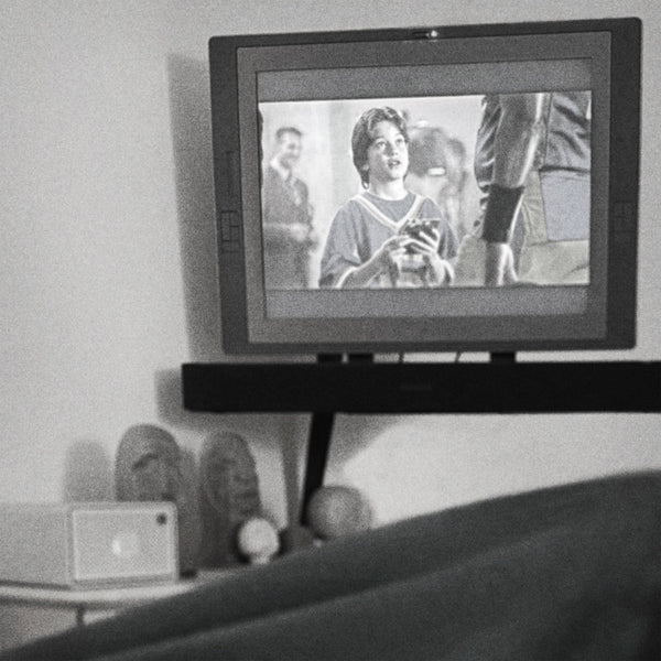 Black-and-white photo taken from under a blanket with feet up, looking at a wall-mounted monitor playing an episode of Psych above a shelf of hand-carved wooden sculptures.