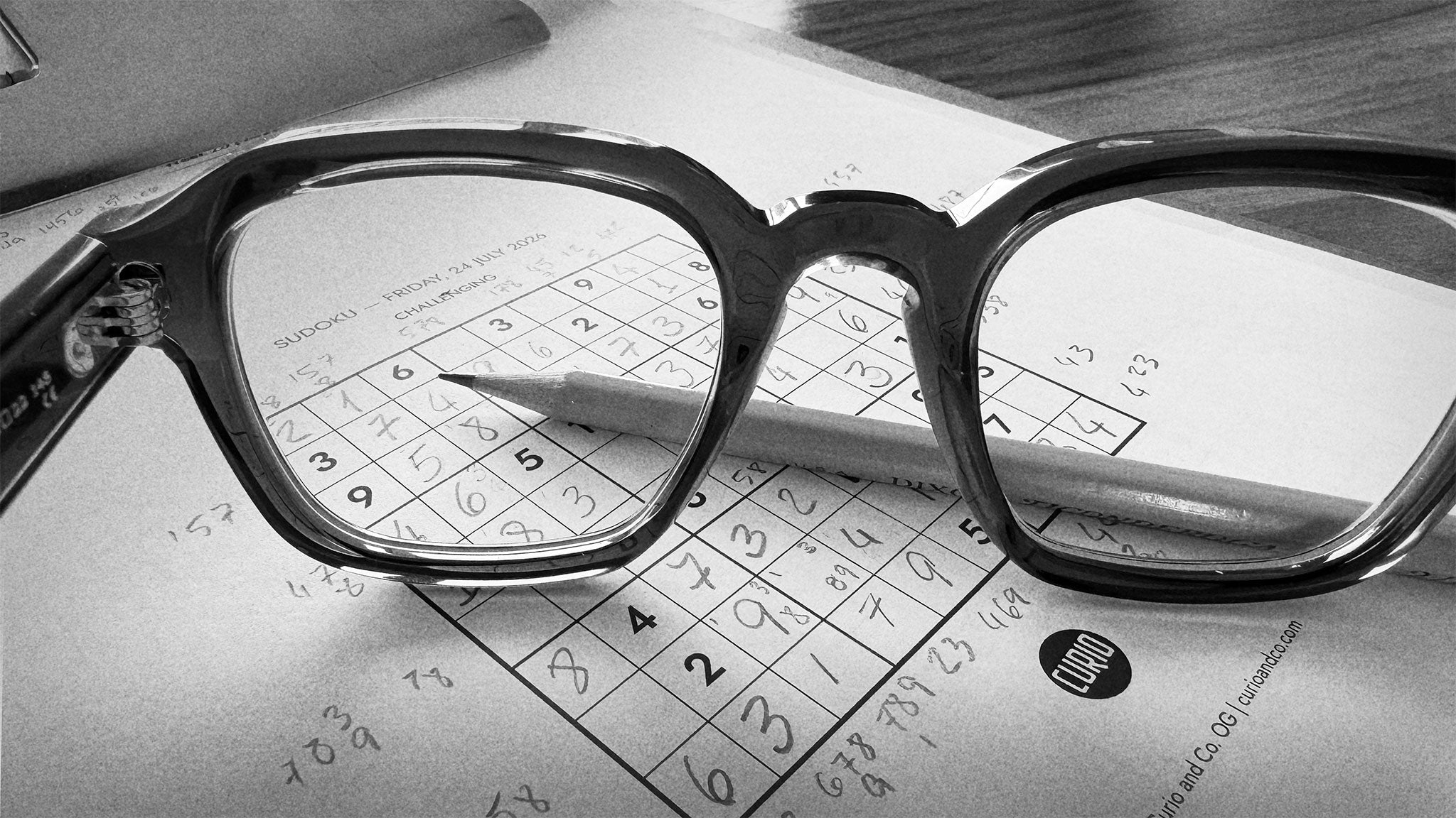 black and white photograph of an almost completed sudoku with pencil an eye glasses laying on it.