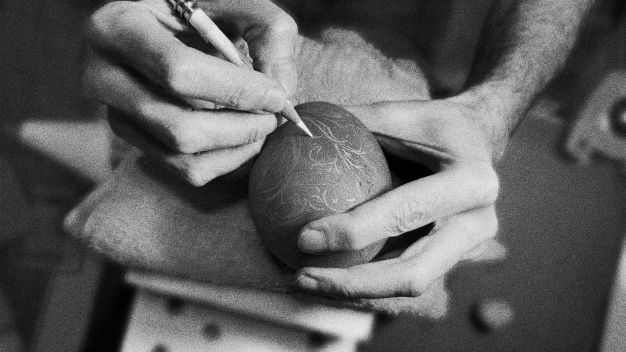 Black & white photo of hands drawing decorative pattern on wood sculpture with white pencil.