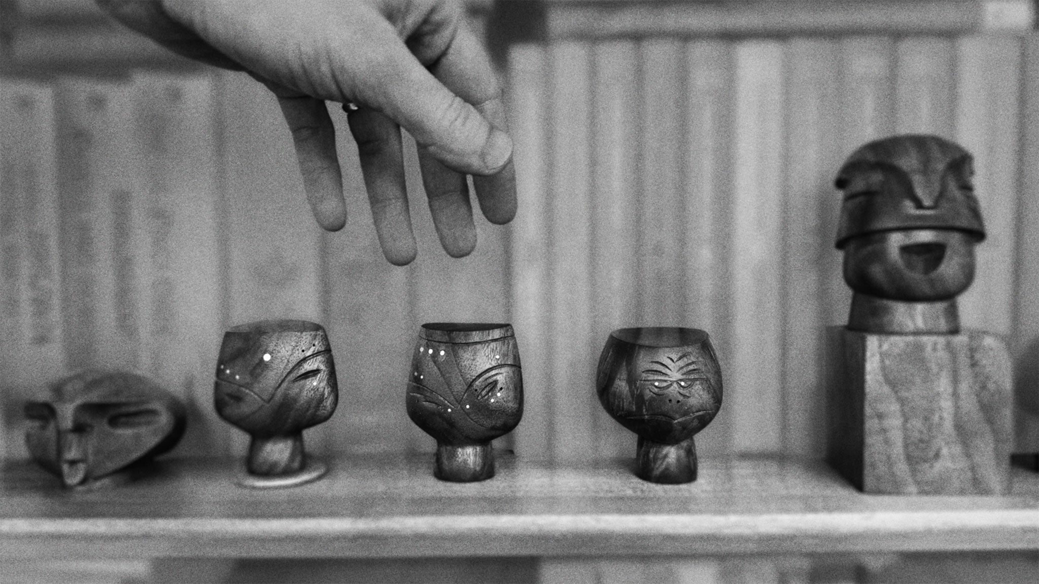 black and white photo o hand reaching for one of 5 mid-century styled wood figures faces on book shelf